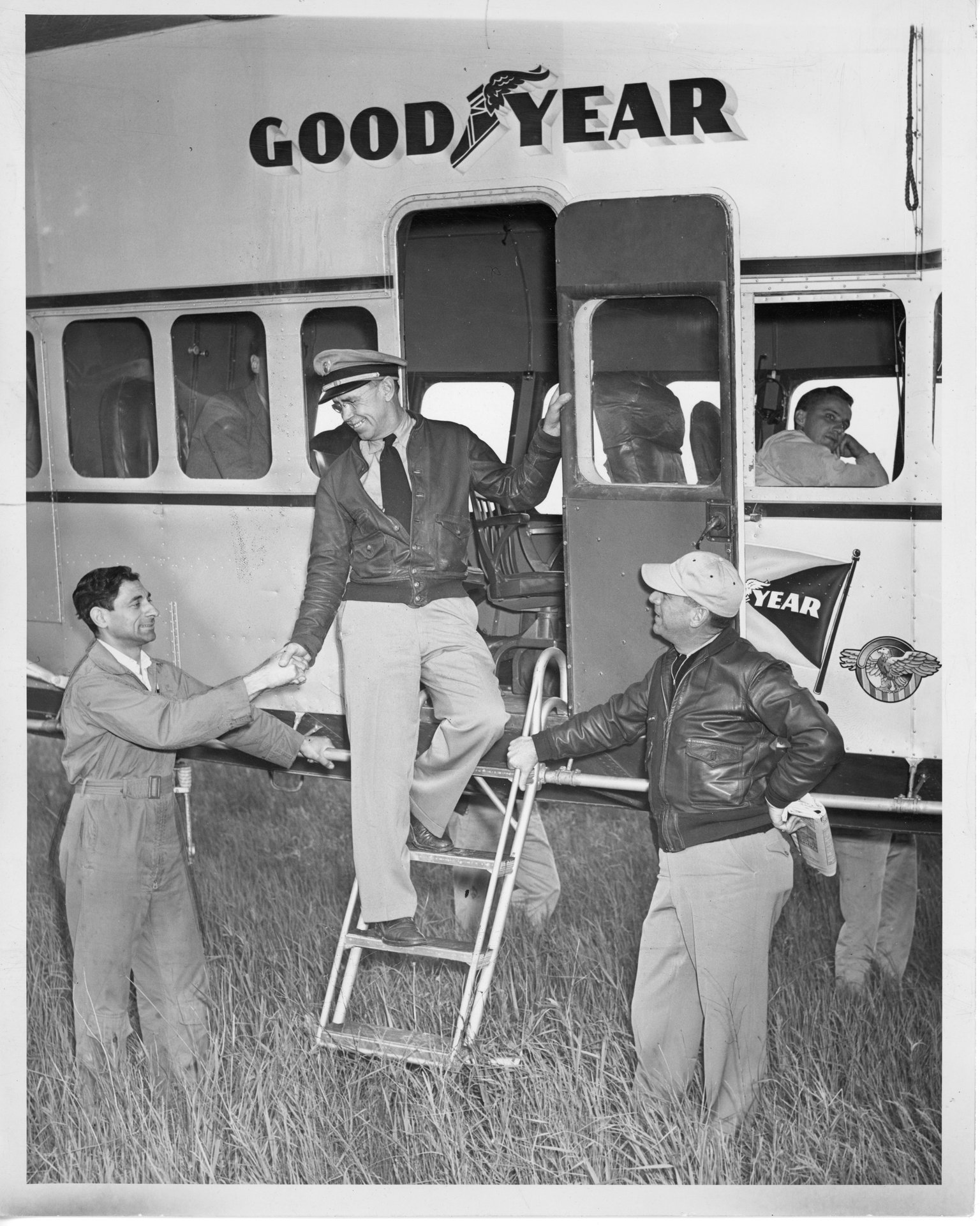 Tony Riccio shaking the Goodyear blimp's pilot's hand with one unknown person standing next to the staircase. Two unknown people are inside the blimp's cabin car.