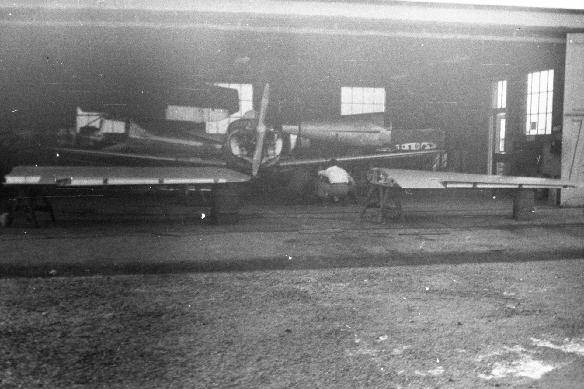 A Globe Swift airplane and separate wings on stands in the Maintenance shop at Buffalo Airpark. Tony Riccio and an unknown person is inspecting the left landing gear of a Globe Swift.