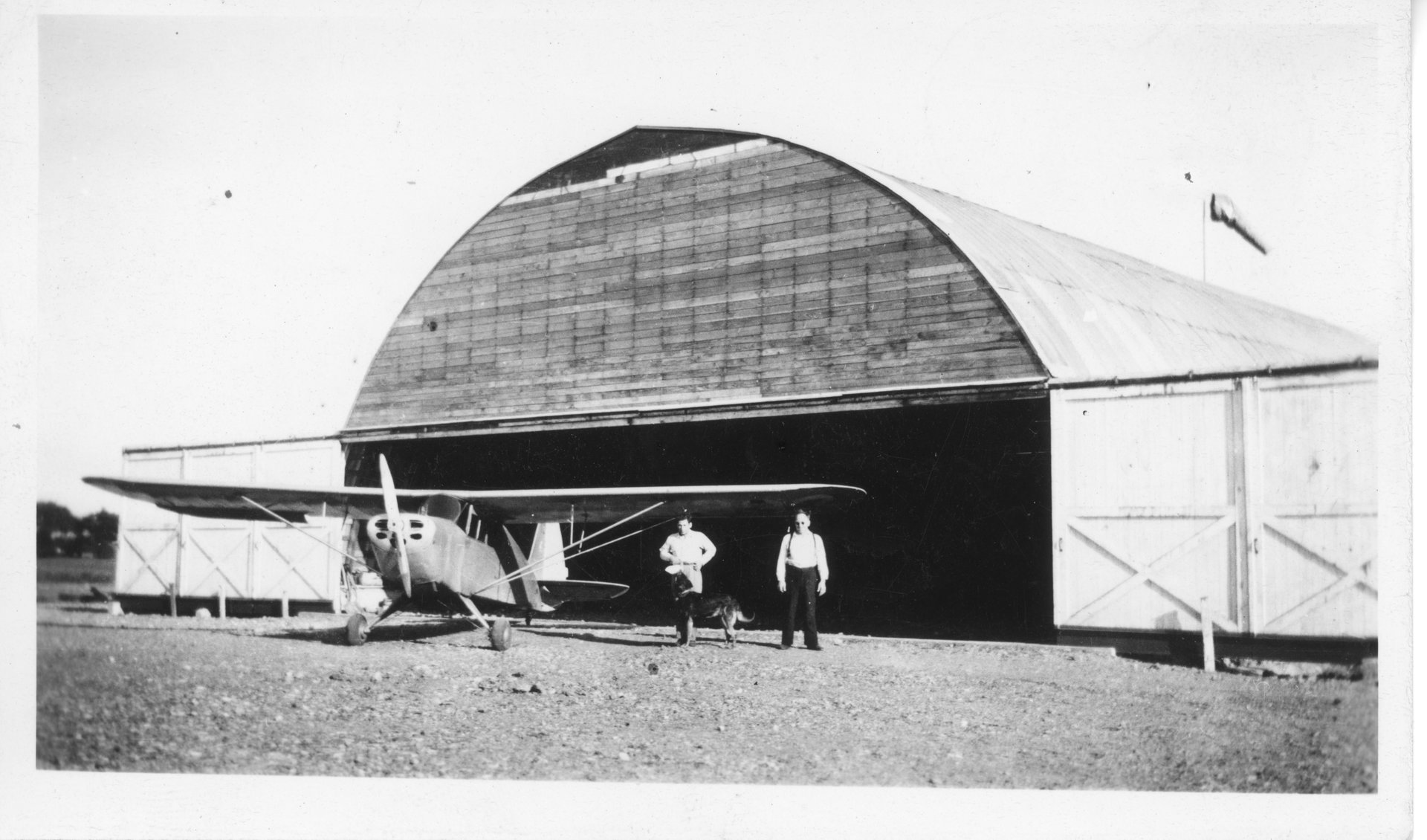 The construction of the Quonset hangar at the Gardenville Airport in August 1944. Tony Riccio, G.B. Baumgarten, and Tony's dog Ivan are standing next to an Interstate S1A F90 airplane.