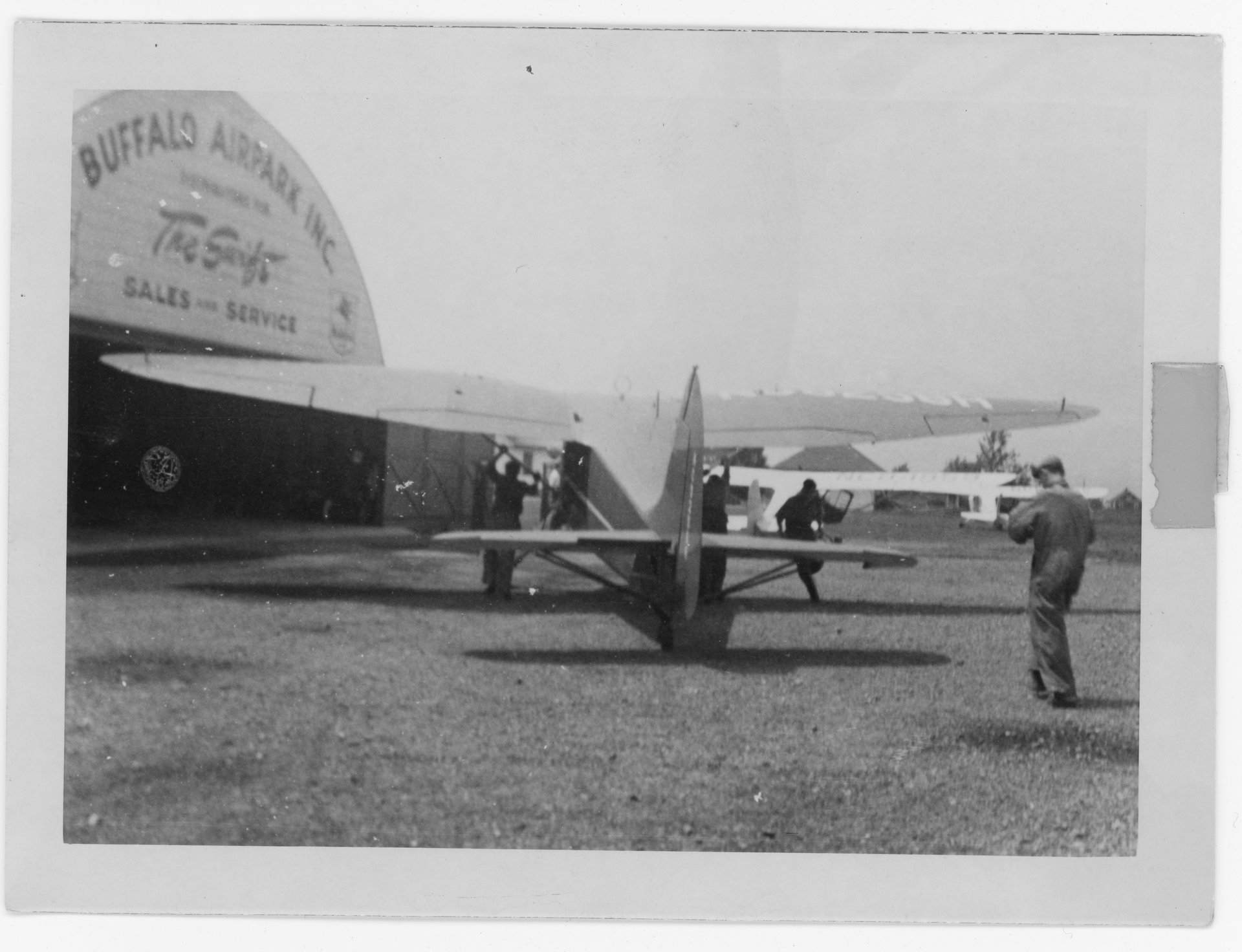 A 1946 Stinson Vultee V-77 is being pushed by three unknown people towards the Quonset hangar at Buffalo Airpark.