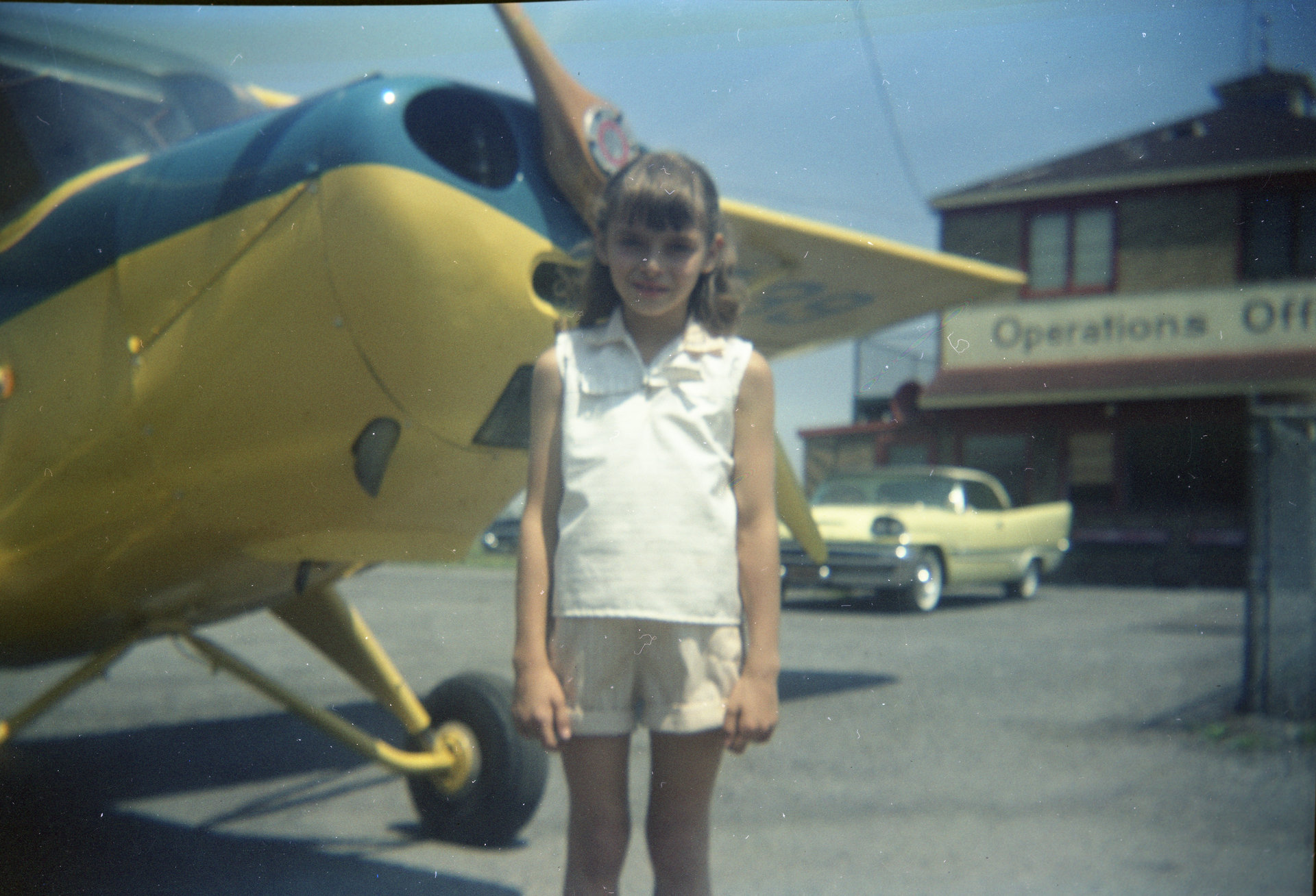 Beverly Riccio, Tony's daughter, standing in front of a yellow Piper airplane with a blue stripe in the parking lot of Buffalo Airpark.