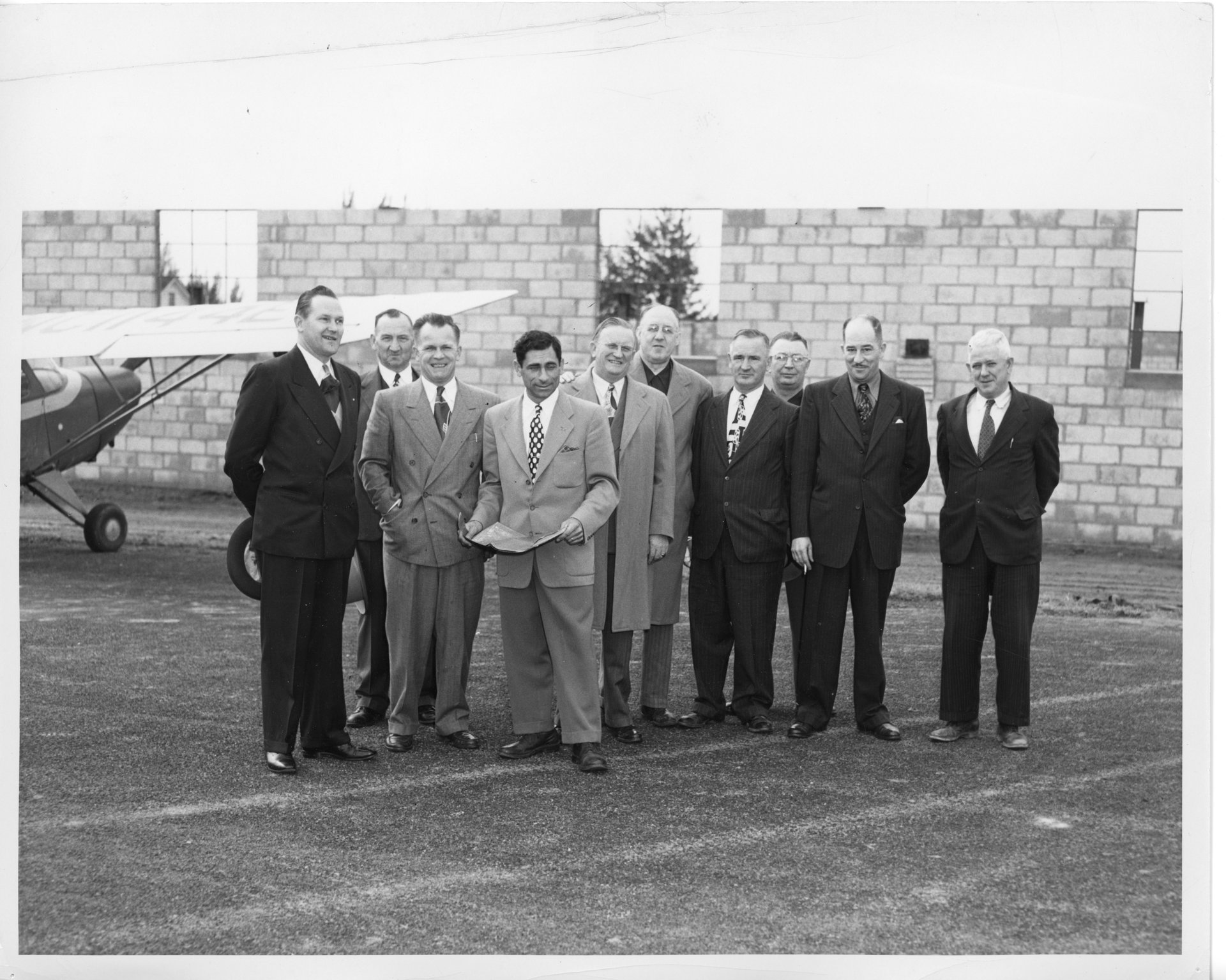 Tony Riccio and nine unknown people grouped together outside of a hangar's brick wall.